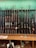 A collection of various rifles is displayed upright against a green felt backing. The rifles have different designs and finishes, featuring wooden and synthetic stocks. Below the rifles, a wooden counter holds miscellaneous items, including a tape measure, some paper, and other small objects.