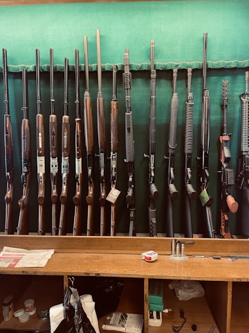 A collection of various rifles is displayed upright against a green felt backing. The rifles have different designs and finishes, featuring wooden and synthetic stocks. Below the rifles, a wooden counter holds miscellaneous items, including a tape measure, some paper, and other small objects.