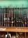 A collection of various rifles is displayed upright against a green felt backing. The rifles have different designs and finishes, featuring wooden and synthetic stocks. Below the rifles, a wooden counter holds miscellaneous items, including a tape measure, some paper, and other small objects.