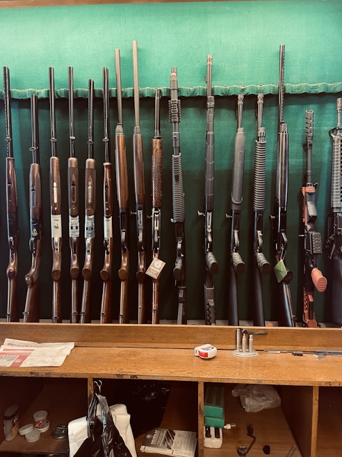 A collection of various rifles is displayed upright against a green felt backing. The rifles have different designs and finishes, featuring wooden and synthetic stocks. Below the rifles, a wooden counter holds miscellaneous items, including a tape measure, some paper, and other small objects.