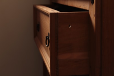 A wooden cabinet with a partially open drawer, featuring a metal ring handle. The wood has a warm, natural finish, and the drawer reveals a shadowed interior. Soft lighting casts gentle shadows, adding depth to the wood grain.