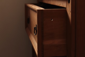 A wooden cabinet with a partially open drawer, featuring a metal ring handle. The wood has a warm, natural finish, and the drawer reveals a shadowed interior. Soft lighting casts gentle shadows, adding depth to the wood grain.