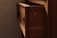 A wooden cabinet with a partially open drawer, featuring a metal ring handle. The wood has a warm, natural finish, and the drawer reveals a shadowed interior. Soft lighting casts gentle shadows, adding depth to the wood grain.