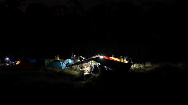 A nighttime camping scene with multiple tents set up on a field, illuminated by small lights. The tents are in various colors including blue and green. There are people gathered around under a shelter, casting a soft glow on the surroundings.