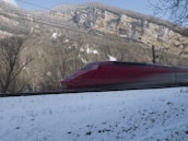 A sleek high-speed train, painted in a bold red color, speeds through a snowy landscape. The train is set against a backdrop of towering mountains and leafless trees, with patches of snow on the ground. Overhead power lines are visible, indicating an electrified railway.