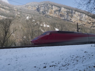 A sleek high-speed train, painted in a bold red color, speeds through a snowy landscape. The train is set against a backdrop of towering mountains and leafless trees, with patches of snow on the ground. Overhead power lines are visible, indicating an electrified railway.