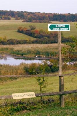 A wooden fence in a grassy field with two signs in German. One sign is mounted on the fence, reading 'WIESE NICHT BETRETEN,' indicating that access to the meadow is restricted. Another green directional sign points to 'RUNDWEG Zentrum Rohrbach.' Behind the fence, a small pond surrounded by reeds is visible, with a backdrop of rolling hills and colorful autumn foliage.