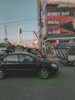 A busy urban intersection with a black car in the foreground and various vehicles, including a rickshaw, parked on the roadside. A tall building displays a large advertisement for a meal with a price, and several people are visible near the vehicles. A mosque minaret is in the background against a clear sky.