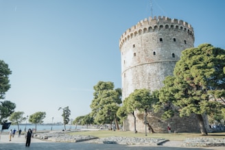 a stone castle with trees and people with White Tower of Thessaloniki in the background