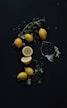A dark background accented with vibrant yellow lemons and green leaves, some lemons whole while others are cut open. A transparent glass juicer is placed nearby, with small white flowers scattered around.