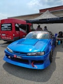 A bright blue racing car is parked in front of a red van at an outdoor event. The car features sponsor decals and modifications typical for race cars. A canopy tent has been set up nearby with people underneath, suggesting a preparation area for a racing event.
