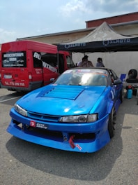 A bright blue racing car is parked in front of a red van at an outdoor event. The car features sponsor decals and modifications typical for race cars. A canopy tent has been set up nearby with people underneath, suggesting a preparation area for a racing event.