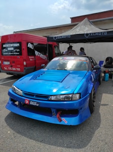 A bright blue racing car is parked in front of a red van at an outdoor event. The car features sponsor decals and modifications typical for race cars. A canopy tent has been set up nearby with people underneath, suggesting a preparation area for a racing event.
