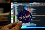 A close-up of a hand holding a circular sticker featuring the NASA logo in front of a computer screen displaying lines of code. The image combines elements of space exploration and technology.
