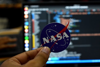 A close-up of a hand holding a circular sticker featuring the NASA logo in front of a computer screen displaying lines of code. The image combines elements of space exploration and technology.