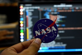 A close-up of a hand holding a circular sticker featuring the NASA logo in front of a computer screen displaying lines of code. The image combines elements of space exploration and technology.