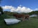 Two brown cows stand near white troughs on a grassy hillside. In the background, there are rolling green hills, a few trees, and a building with a solar-paneled roof under a blue sky filled with fluffy white clouds. A tractor is partially visible on the left side, and a wire fence separates the cows from the field.