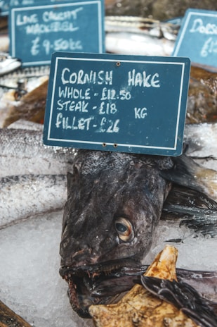An assortment of fresh fish on ice, displayed at a market. A sign prominently advertises Cornish hake with prices for whole, steak, and fillet portions. Other fish varieties are partially visible in the background with additional signs showing prices.