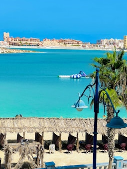 A serene coastal scene with bright turquoise waters and a clear blue sky. Straw umbrellas line the sandy beach, offering shade, while a few plastic chairs are scattered beneath them. In the distance, modern buildings rise along the shoreline, and a large inflatable structure floats on the calm water. A palm tree and a decorative lamppost frame the foreground.