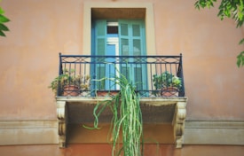 A small rustic balcony with decorative black metal railings and potted plants, situated against a warm, peach-colored wall. The closed wooden shutters of the window are painted in a light shade of green. There are lush, trailing plants that hang gracefully over the edge of the balcony.