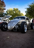 A custom, vintage car with a shiny, metallic finish is parked on a gravel surface. The car has a unique, sleek design with large, exposed wheels and a lowered body. In the background, there are trees with dense green foliage and a partly cloudy blue sky. Another vehicle with its trunk open is partially visible.