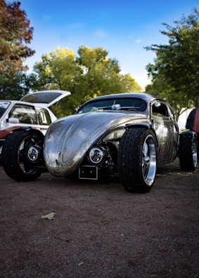 A custom, vintage car with a shiny, metallic finish is parked on a gravel surface. The car has a unique, sleek design with large, exposed wheels and a lowered body. In the background, there are trees with dense green foliage and a partly cloudy blue sky. Another vehicle with its trunk open is partially visible.