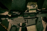 A close-up view of a firearm, specifically an assault rifle, showing detailed components such as the safety switch and various mounted accessories. The rifle is resting on a wooden surface.