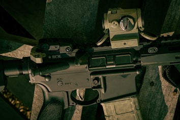 A close-up view of a firearm, specifically an assault rifle, showing detailed components such as the safety switch and various mounted accessories. The rifle is resting on a wooden surface.