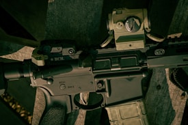 A close-up view of a firearm, specifically an assault rifle, showing detailed components such as the safety switch and various mounted accessories. The rifle is resting on a wooden surface.