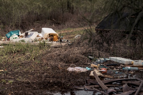 A cluttered arrangement of trash and debris sitting among tall grass and muddy ground. Various items, including plastic bottles, cardboard, and other garbage, are scattered across the area. The scene is untidy and highlights a lack of cleanliness in a natural environment.