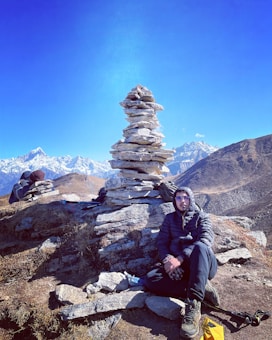 A person dressed in outdoor gear is sitting beside a tall stack of balanced rocks on a rugged mountain landscape. The background features snow-capped peaks under a clear blue sky. Another individual appears to be resting on the other side of the rock pile.