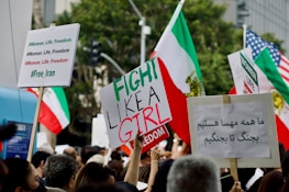 A large group of people holding various protest signs in different languages, adorned with hashtags like #Woman_Life_Freedom and #Free_Iran. The scene is filled with Iranian flags and some American flags, suggesting a protest or demonstration focused on issues surrounding Iran and women's rights, likely in an urban setting judging by the background.