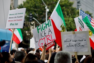 A large group of people holding various protest signs in different languages, adorned with hashtags like #Woman_Life_Freedom and #Free_Iran. The scene is filled with Iranian flags and some American flags, suggesting a protest or demonstration focused on issues surrounding Iran and women's rights, likely in an urban setting judging by the background.