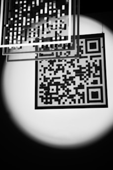 A black and white image features a QR code illuminated by a soft spotlight. Several transparent squares with grid patterns are suspended above, adding a layered, abstract effect. The contrast between light and dark creates a striking, almost mysterious atmosphere.