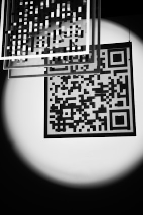 A black and white image features a QR code illuminated by a soft spotlight. Several transparent squares with grid patterns are suspended above, adding a layered, abstract effect. The contrast between light and dark creates a striking, almost mysterious atmosphere.