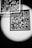 A black and white image features a QR code illuminated by a soft spotlight. Several transparent squares with grid patterns are suspended above, adding a layered, abstract effect. The contrast between light and dark creates a striking, almost mysterious atmosphere.