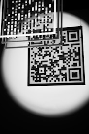 A black and white image features a QR code illuminated by a soft spotlight. Several transparent squares with grid patterns are suspended above, adding a layered, abstract effect. The contrast between light and dark creates a striking, almost mysterious atmosphere.