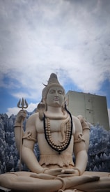 A large statue depicting a figure seated in a meditative pose, holding a trident in the right hand. The statue is adorned with necklaces and has long, stylized hair. A small building is visible in the background against a backdrop of a cloudy sky.