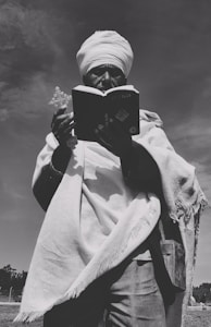 A person wearing traditional clothing and a turban stands outdoors, holding a book and a cross in their hands. The image is in black and white, creating a timeless and contemplative atmosphere. The person appears to be engaged in an act of reading or prayer, with a clear sky in the background.
