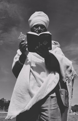 A person wearing traditional clothing and a turban stands outdoors, holding a book and a cross in their hands. The image is in black and white, creating a timeless and contemplative atmosphere. The person appears to be engaged in an act of reading or prayer, with a clear sky in the background.