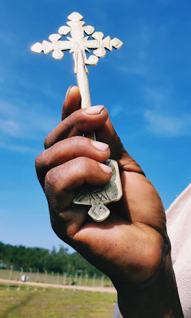 A close-up of a hand holding a small wooden cross against a backdrop of sandy dunes.
