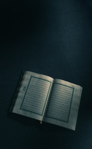 An open book with Arabic script, resting on a dimly lit surface. The pages show intricate designs and bordered text, suggesting religious or traditional content.