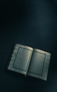 An open book with Arabic script, resting on a dimly lit surface. The pages show intricate designs and bordered text, suggesting religious or traditional content.