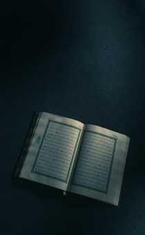 An open book with Arabic script, resting on a dimly lit surface. The pages show intricate designs and bordered text, suggesting religious or traditional content.