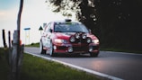 A red rally car speeds down a narrow road, equipped with multiple large headlights on the front bumper. The car's headlights are brightly lit, and sponsor logos are visible along the body. The road is bordered by grass and trees, with road signs in the background.