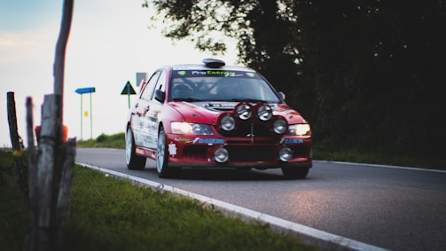 A red rally car speeds down a narrow road, equipped with multiple large headlights on the front bumper. The car's headlights are brightly lit, and sponsor logos are visible along the body. The road is bordered by grass and trees, with road signs in the background.