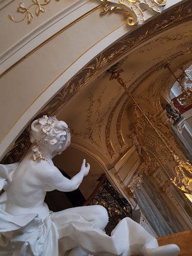 An elegant white marble statue is positioned beneath a grand, ornate archway with intricate gold detailing and carvings. The statue depicts a figure in flowing attire with exquisite craftsmanship. Above, the lavish ceiling features elaborate designs and chandeliers, contributing to the opulent atmosphere.