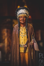 A wooden statue of a Native American man, dressed in traditional attire with a feather headdress, a yellow shirt, and a brown cloak. He is holding a small American flag in his right hand and has several beaded necklaces. The statue is placed indoors and the background is dark and blurred, highlighting the statue distinctly.