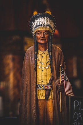 A wooden statue of a Native American man, dressed in traditional attire with a feather headdress, a yellow shirt, and a brown cloak. He is holding a small American flag in his right hand and has several beaded necklaces. The statue is placed indoors and the background is dark and blurred, highlighting the statue distinctly.