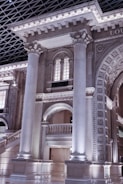 A grand architectural interior featuring large classical columns and intricately detailed arches made of stone. The structure includes ornate designs and a high ceiling with a geometric pattern. Soft lighting highlights the elaborate carvings and intricate moldings on the pillars and arches.